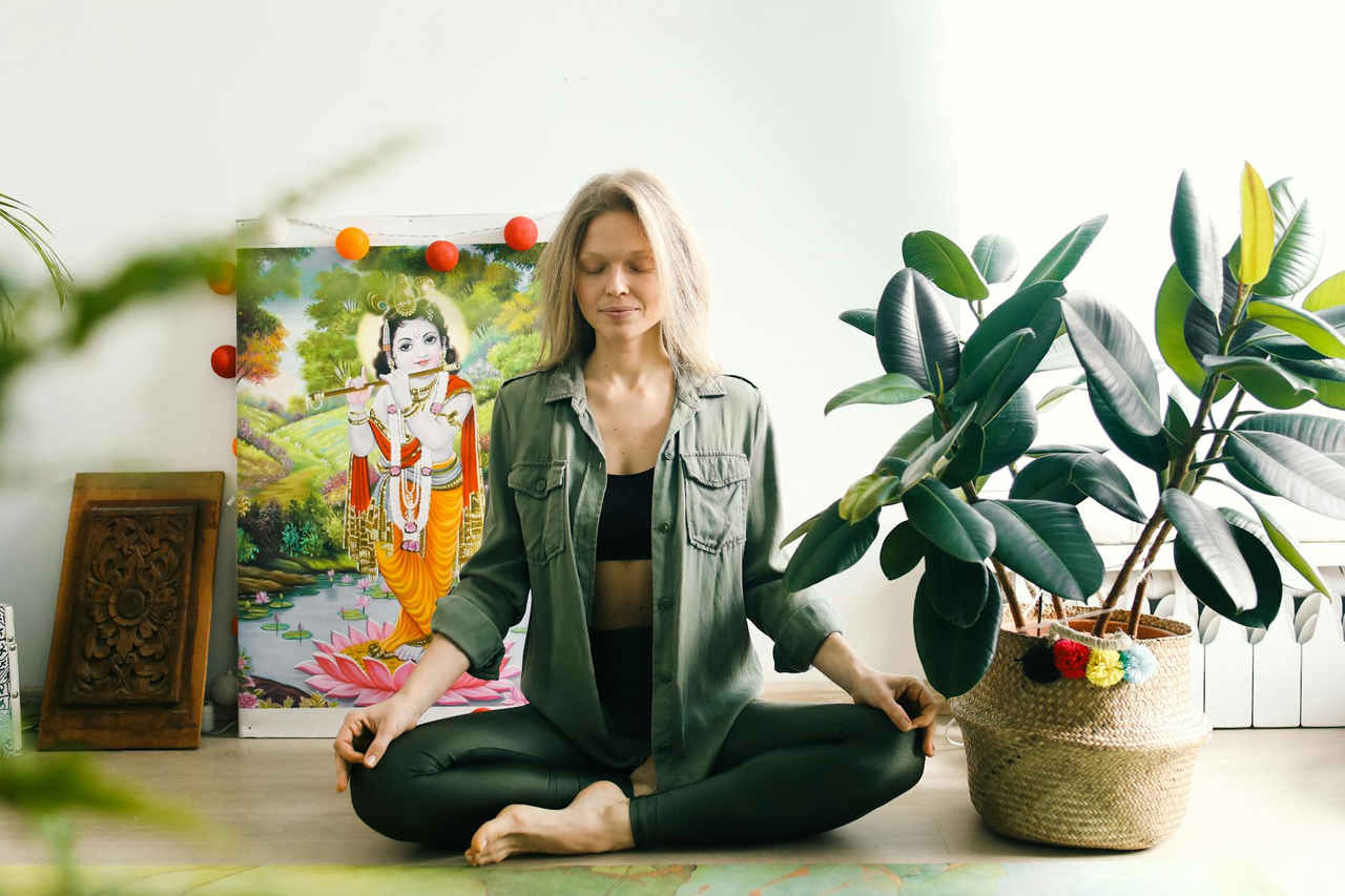 Woman meditating indoors with closed eyes, surrounded by vibrant plants and spiritual art for wellness.